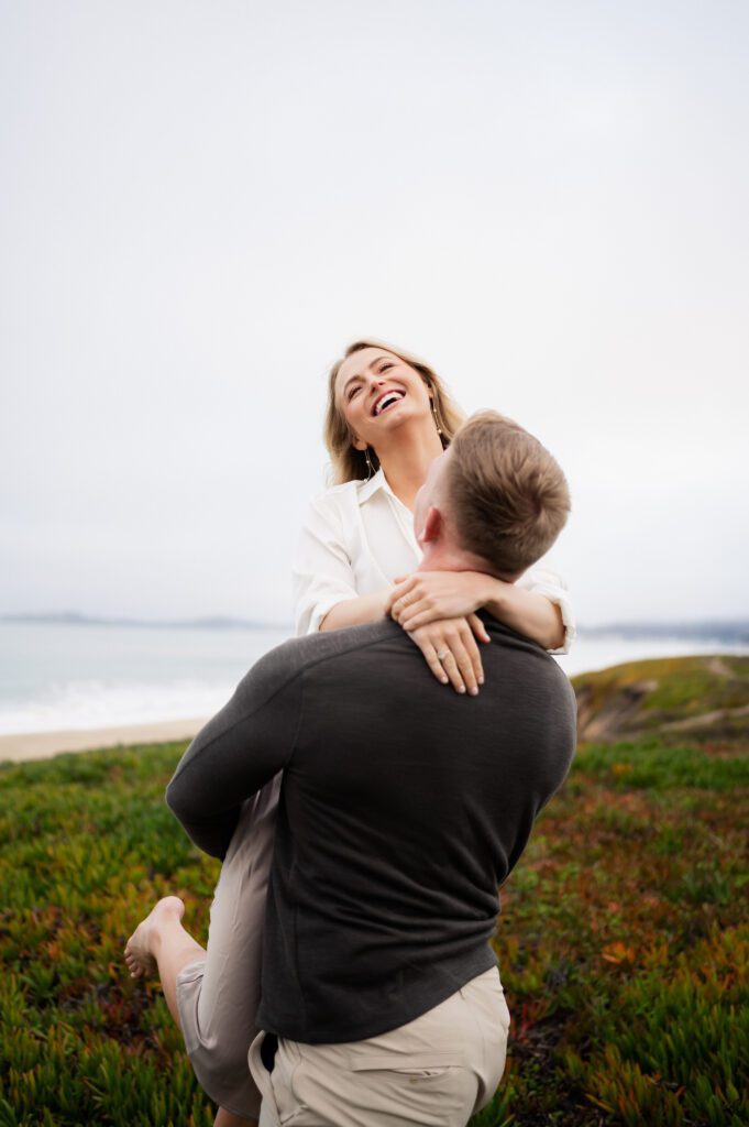 Man picks up and twirls his fiance during half moon bay engagements with san francisco, california wedding photographer and videographer team.