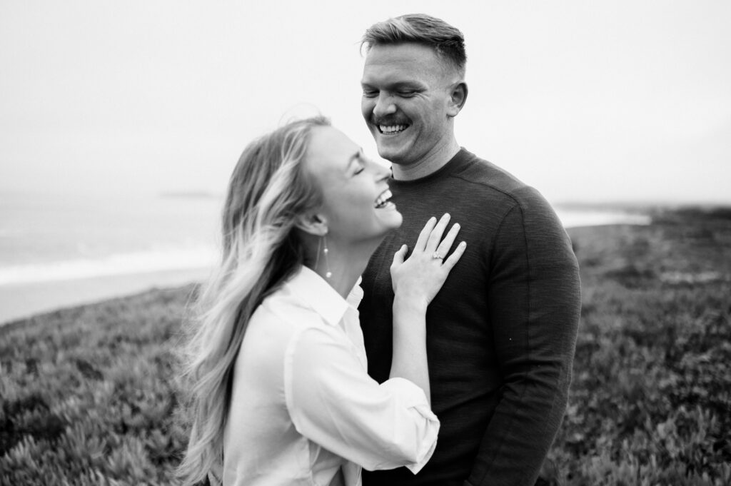 Engaged couple laughs together casually during half moon bay engagements with san francisco, california wedding photographer and videographer team.