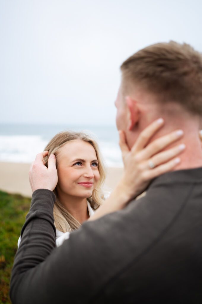 Man tucks his fiance's hair behind her ear during half moon bay engagements with san francisco, california wedding photographer and videographer team.